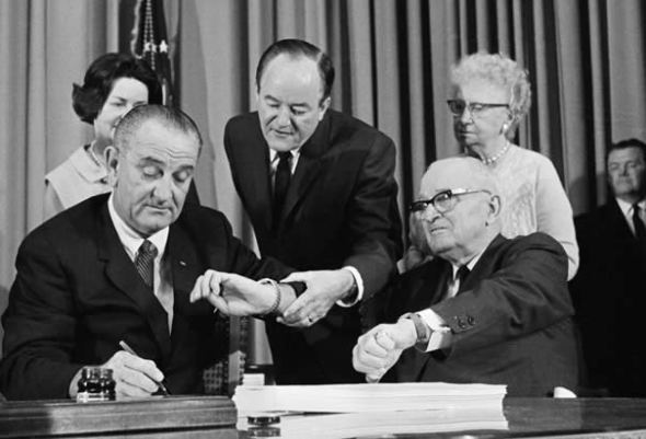 Lyndon B. Johnson signs the Medicare Act of 1964, with Harry Truman by his side