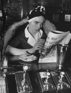 A woman wearing a turban while drinking a chocolate shake and reading the newspaper